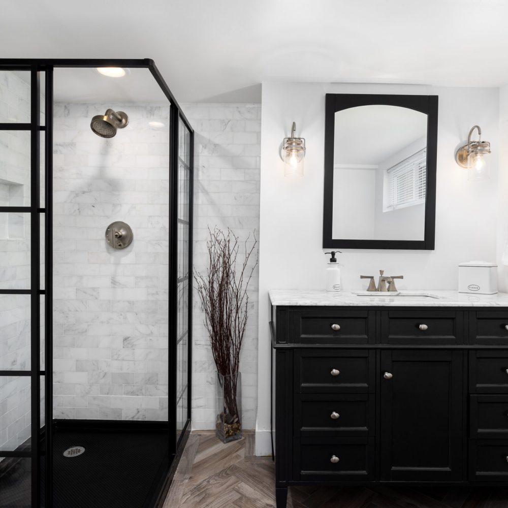A bathroom with white walls, black cabinet and shower, marble subway tiles, and bronze sconces and faucet.