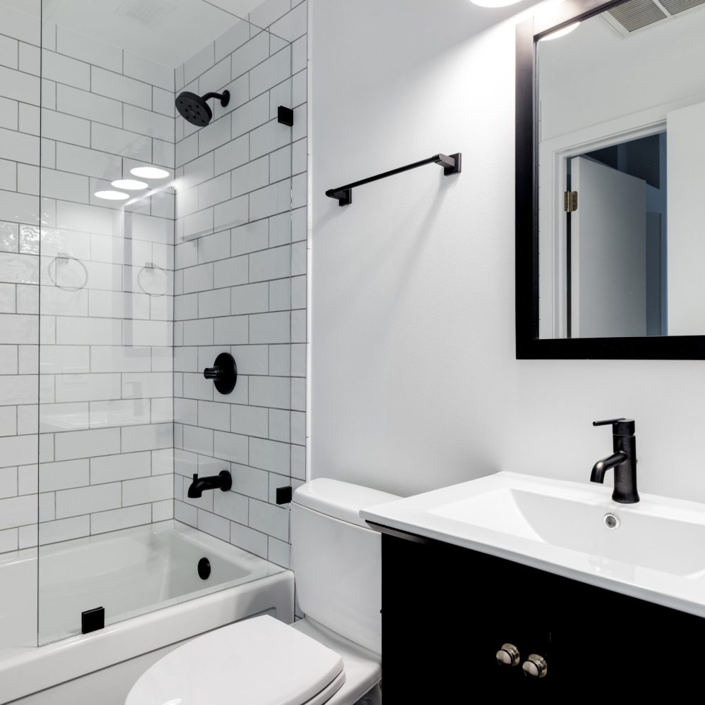 A small modern bathroom with a dark vanity, mirror frame, and hardware. White subway tiles line the bathtub and shower with black faucets.