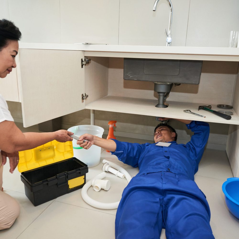 Plumber asking senior woman to give him wrench when he is repairing leaking pipe